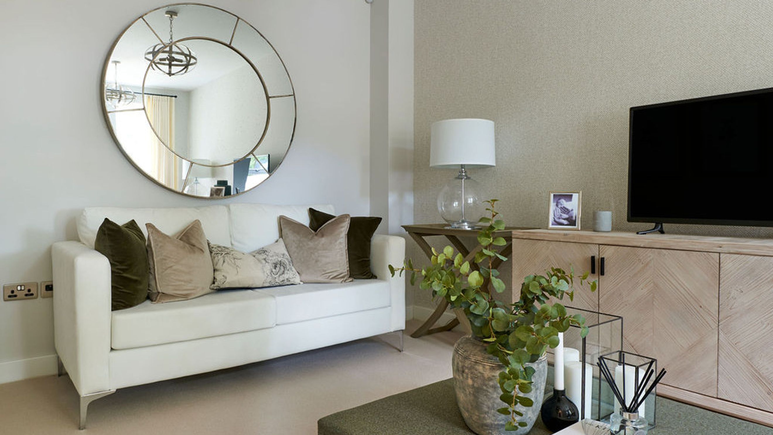 A living room with a white sofa, a large round mirror, a television, and a media console.