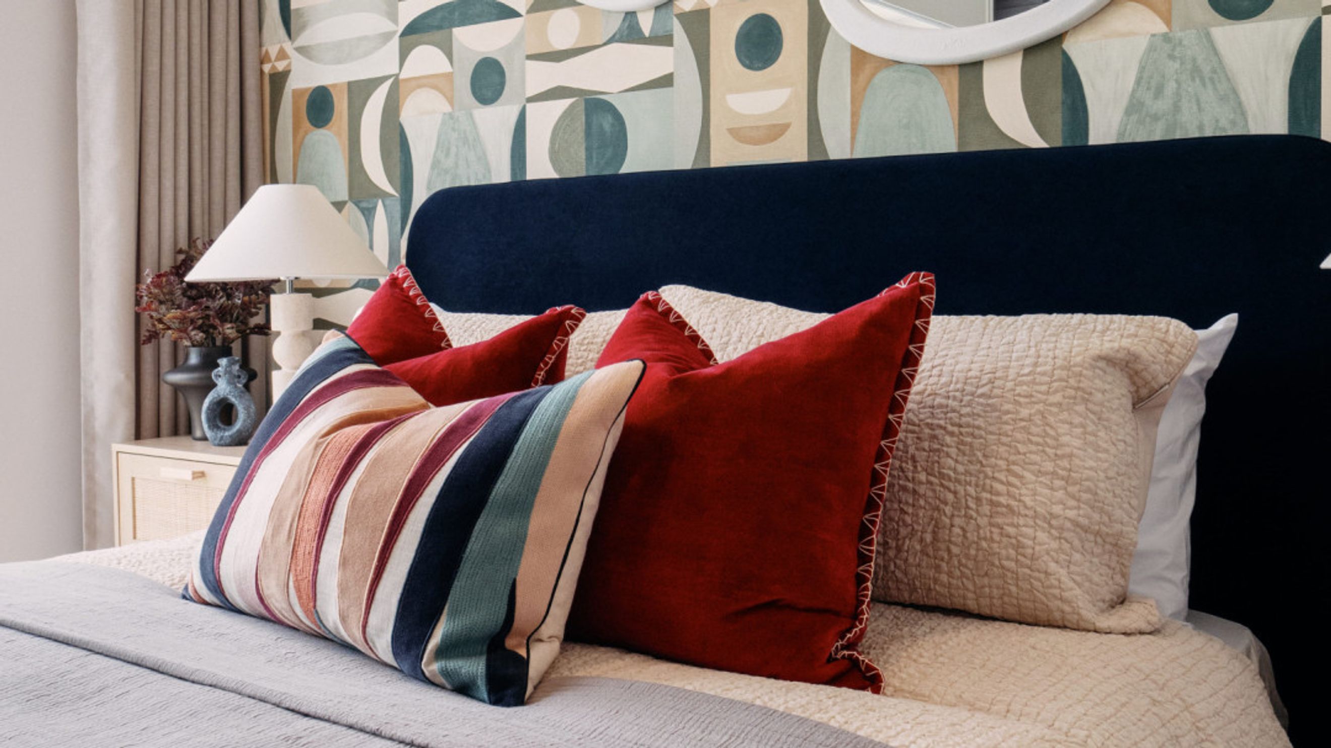 A bedroom with a bed, pillows, a headboard, a lamp, and wallpaper.