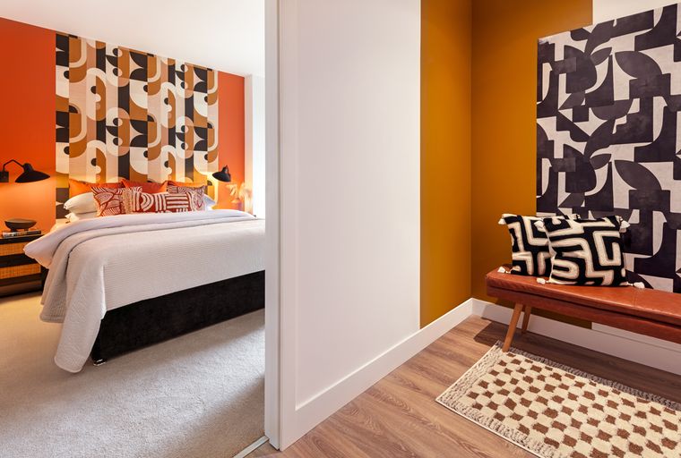 A bedroom with a bed and a geometric accent wall, viewed from a hallway with a bench and pillows.
