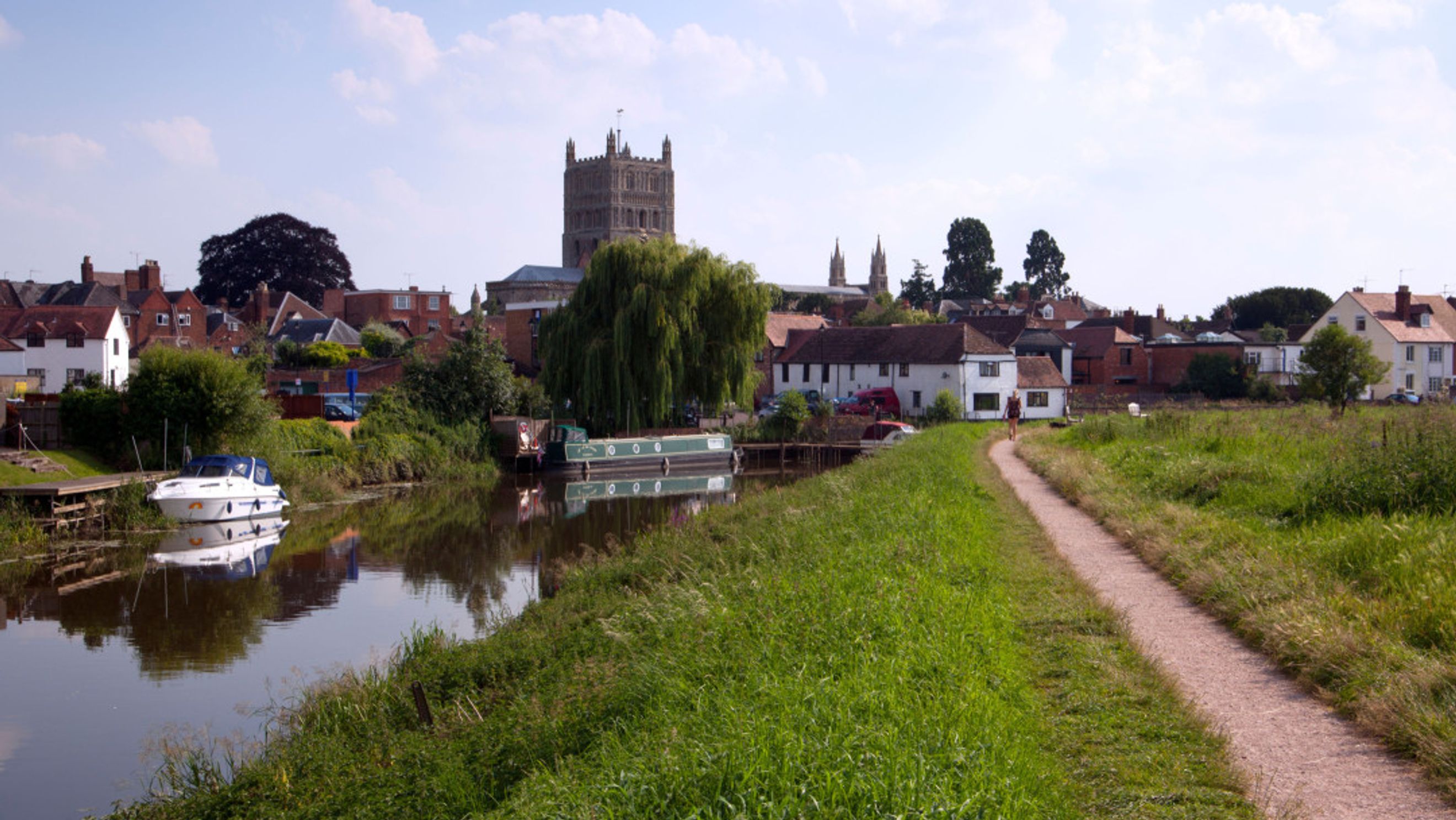 A riverfront view of a town with a cathedral, boats on the water, and a walking path.