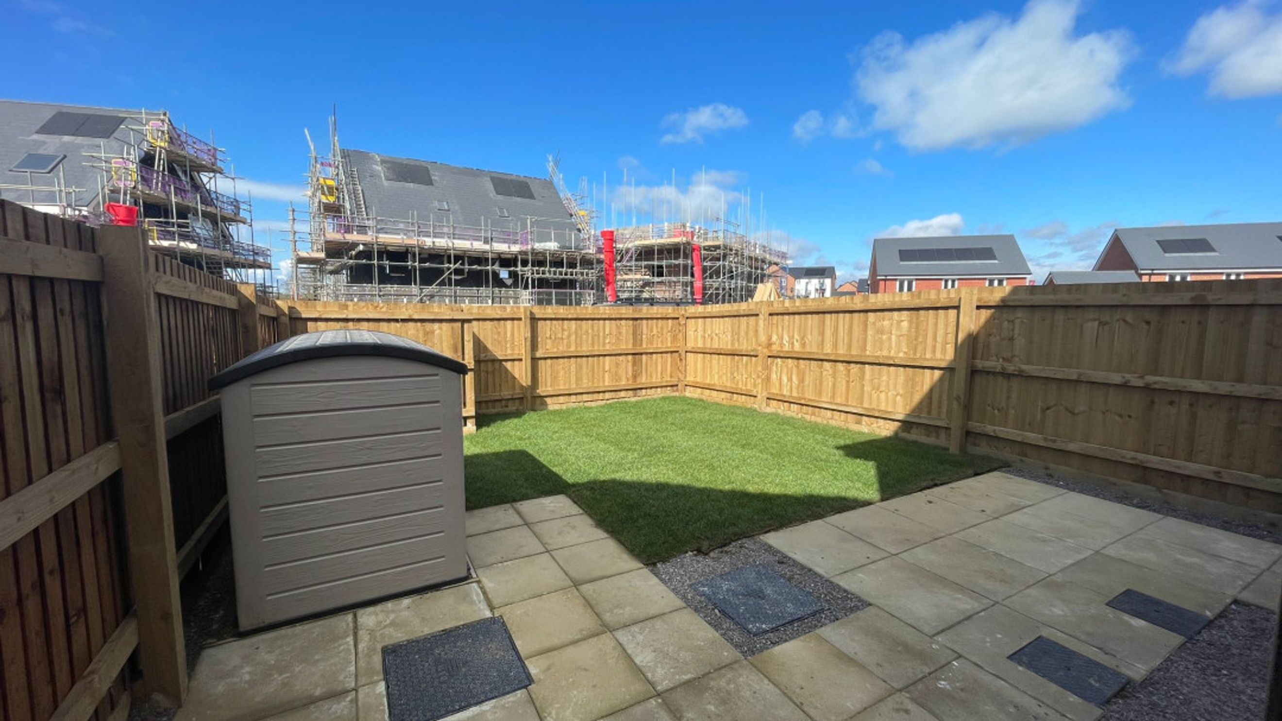 A backyard with a lawn, patio, storage shed, and wooden fence, with houses under construction in the background.