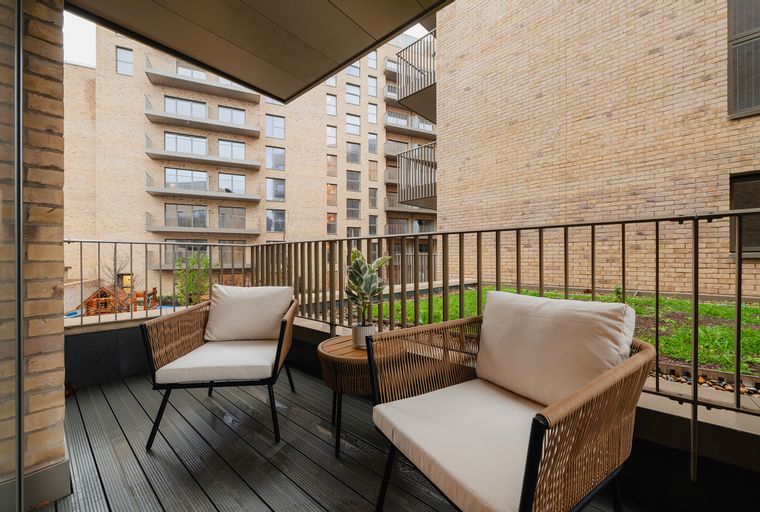 A balcony with two chairs, a small table, and a potted plant, overlooking a courtyard with grass and modern buildings.