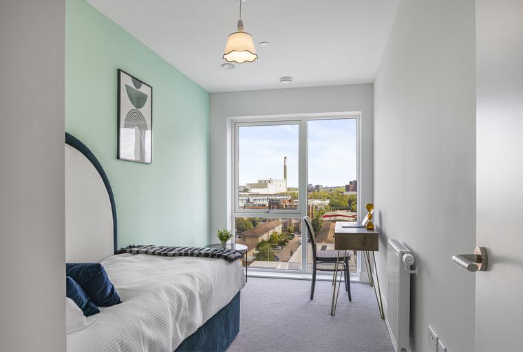 A bedroom with a bed, a desk, a chair, and a large window overlooking a cityscape.