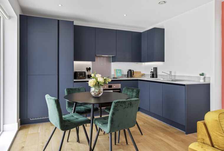 A modern kitchen with dark blue cabinets, a round dining table, and green velvet chairs.