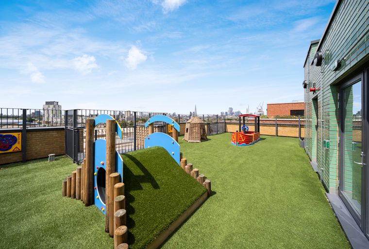 A rooftop playground with artificial turf and various play structures, overlooking a city skyline.