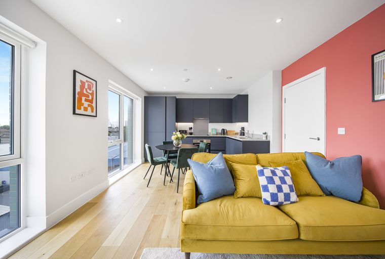A living room and kitchen area with a yellow sofa, dining table, and dark cabinets.