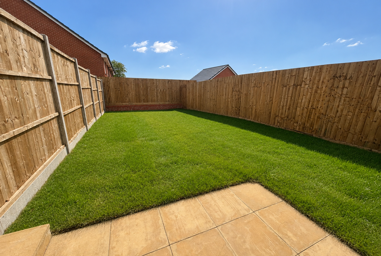 A backyard with a lawn, a patio, and a wooden fence under a blue sky.
