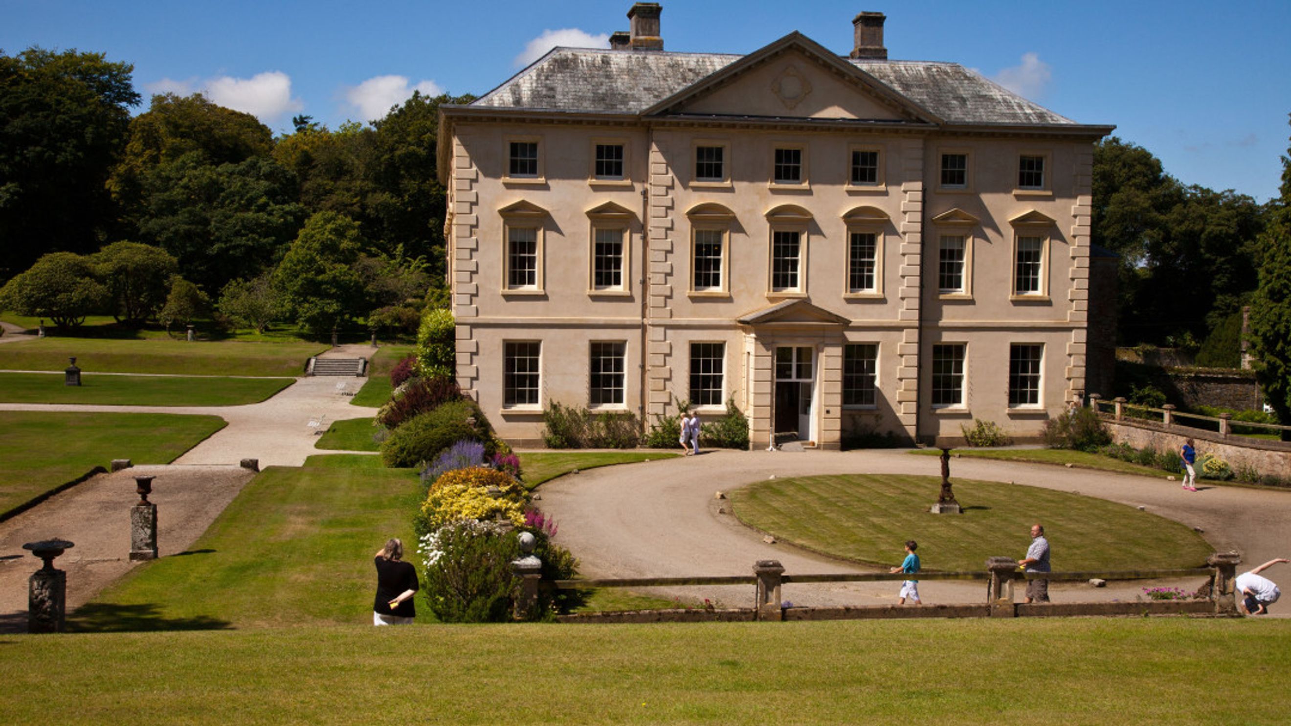 A large, light-colored mansion with many windows is surrounded by a green lawn and trees.