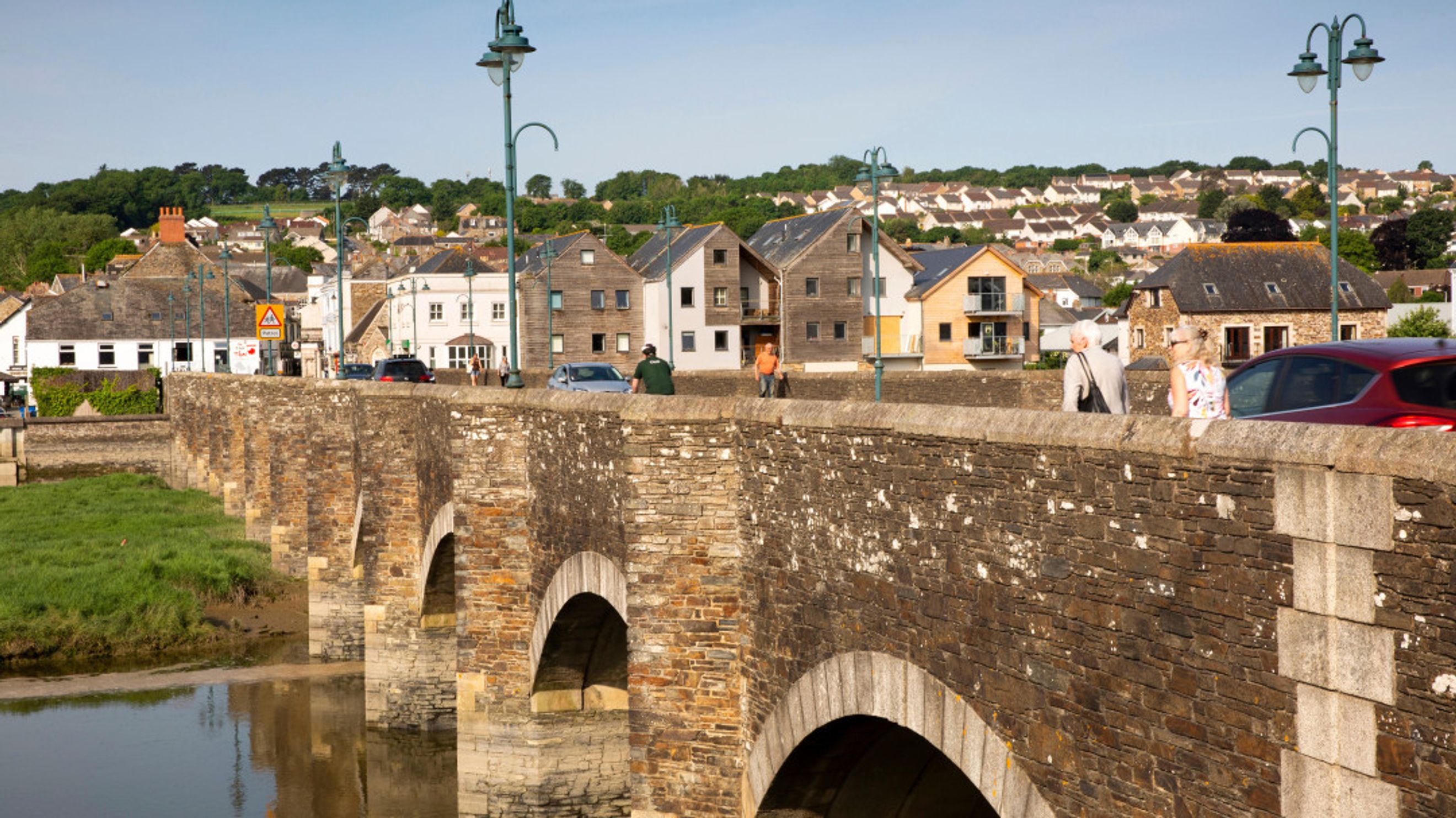 A stone bridge with arches crosses a body of water, with a town and houses in the background.