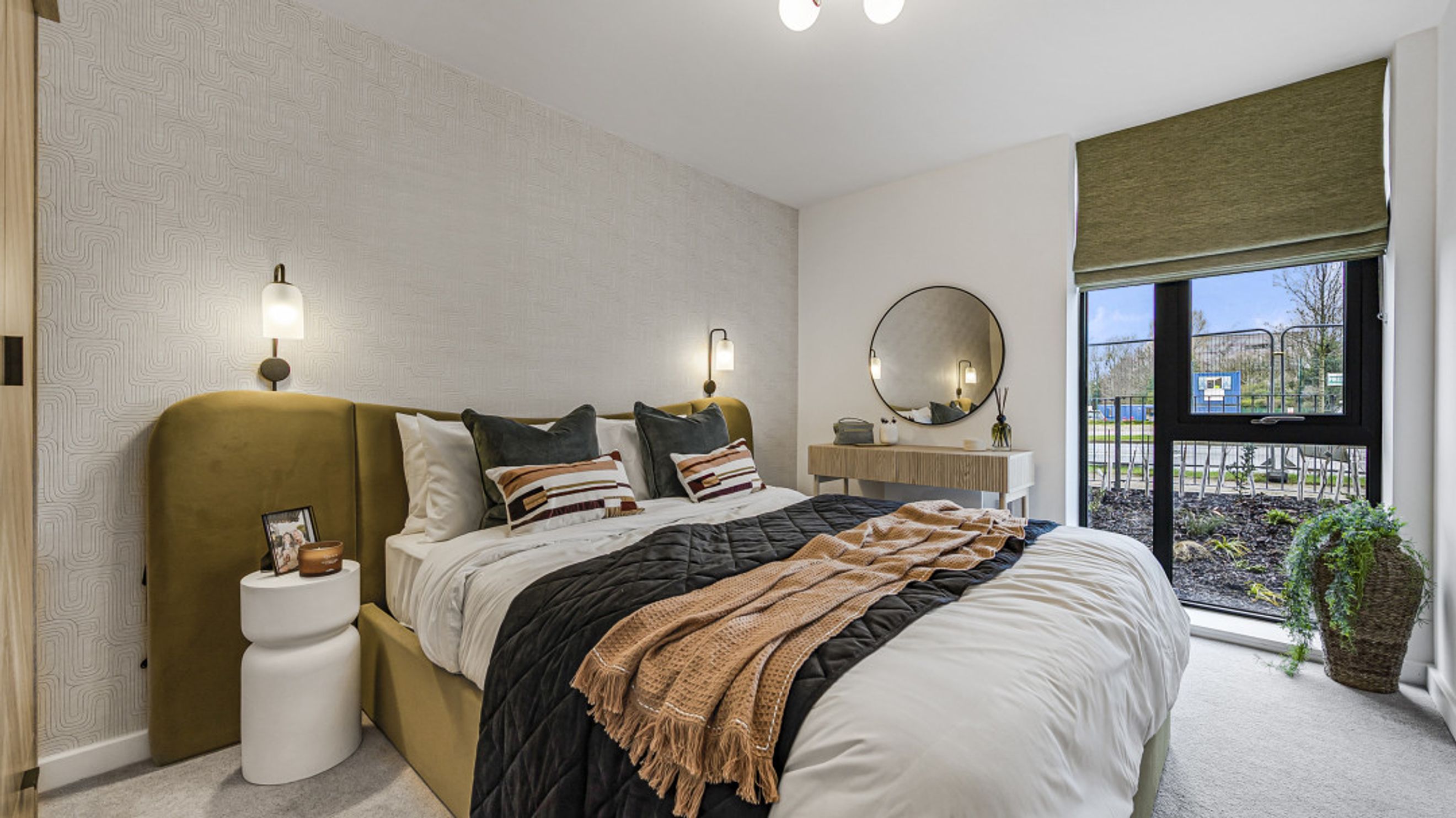 A bedroom with a bed, pillows, a throw blanket, a bedside table, a vanity, a mirror, and a window.
