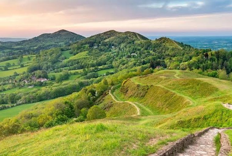 A view of rolling green hills with walking paths and trees under a cloudy sky.