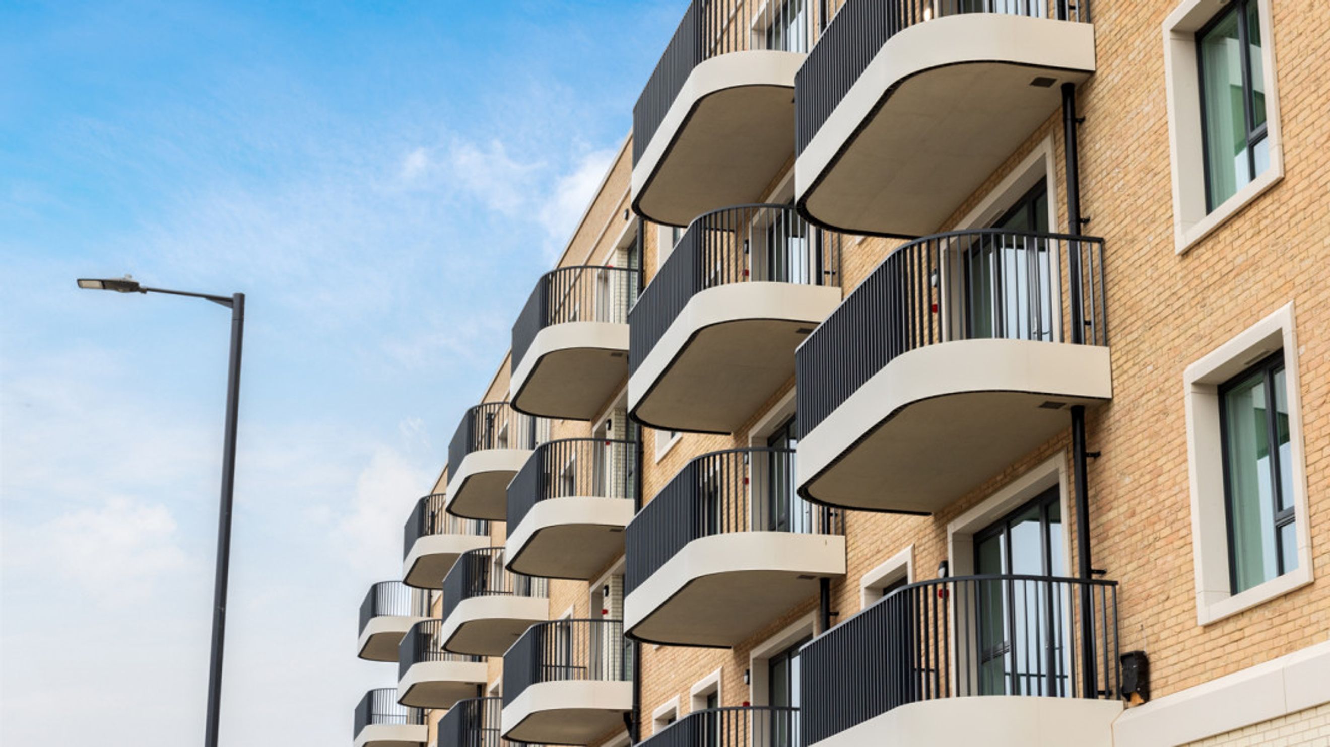 A modern apartment building with curved balconies and light brown brick exterior under a blue sky.