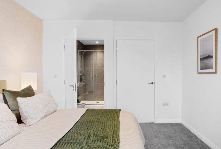 A bedroom with a bed, pillows, a blanket, a lamp, a doorway to a shower, and a framed picture on the wall.