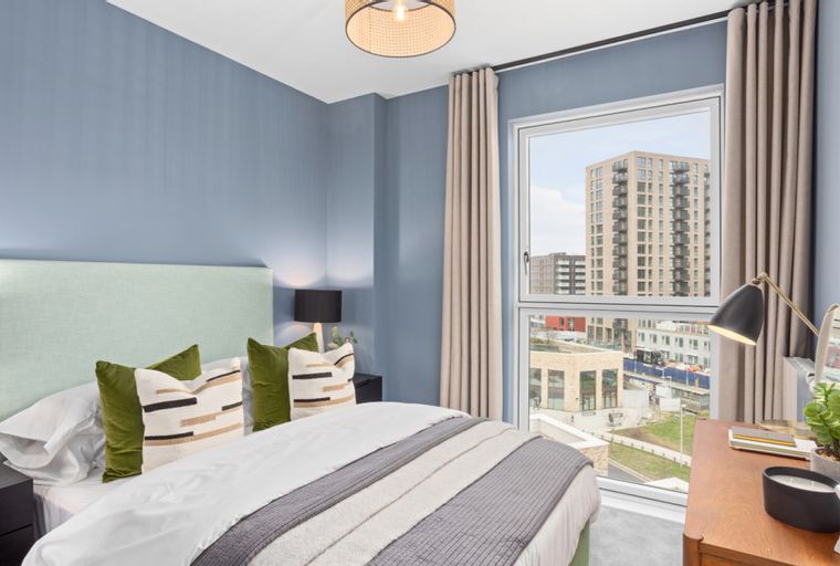 A bedroom with a bed, pillows, a nightstand, a desk, and a window overlooking a city.