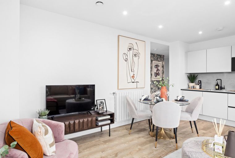 A living and dining area with a kitchen, featuring a pink armchair, a TV, a dining table with chairs, and white kitchen cabinets.