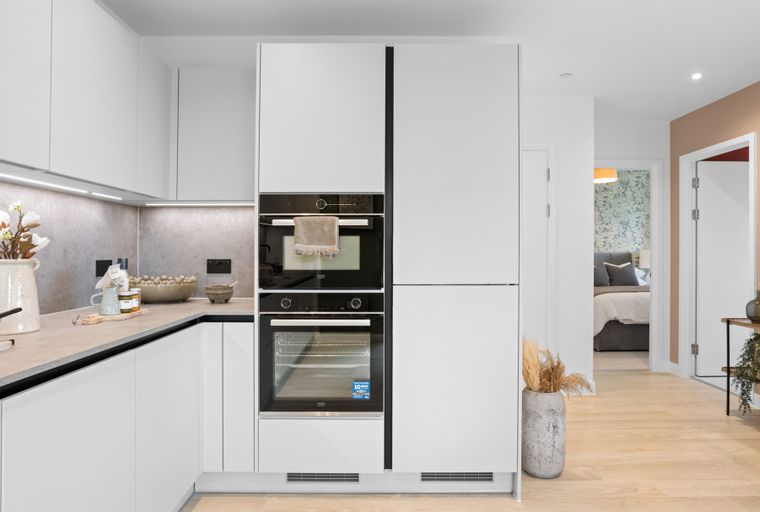 A modern kitchen with white cabinets, integrated ovens, and a countertop, leading to a hallway with a bedroom and entryway.