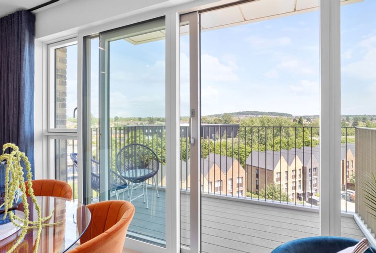 A dining area with orange chairs and a glass table next to a sliding glass door leading to a balcony with a view of houses and trees.