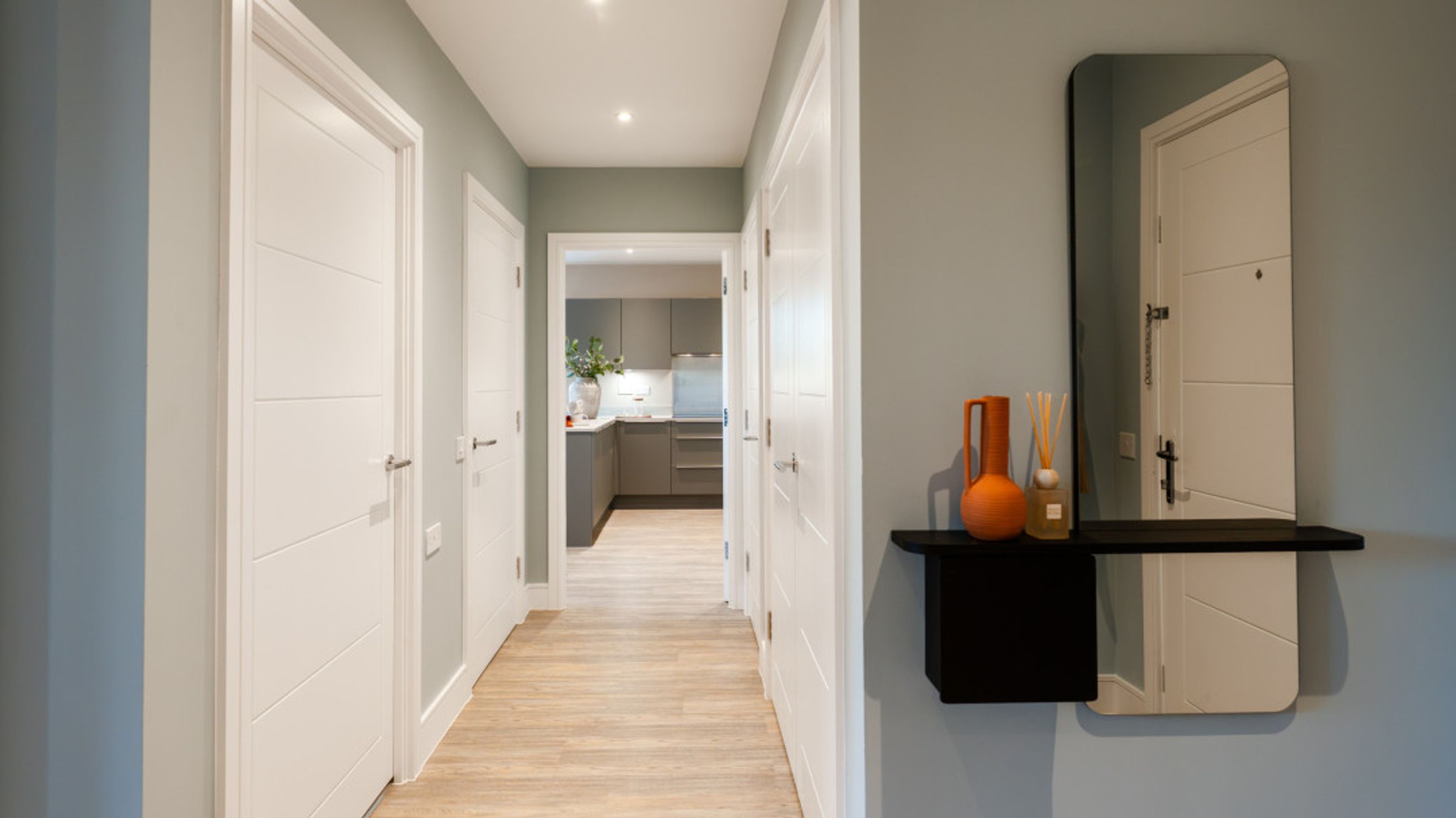A hallway with white doors, a wood-effect floor, and a view into a kitchen.