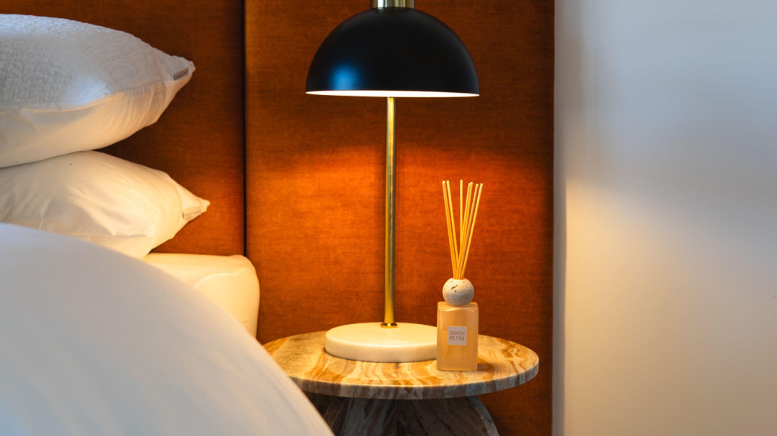 A bedside table with a lamp and a reed diffuser next to a stack of pillows.