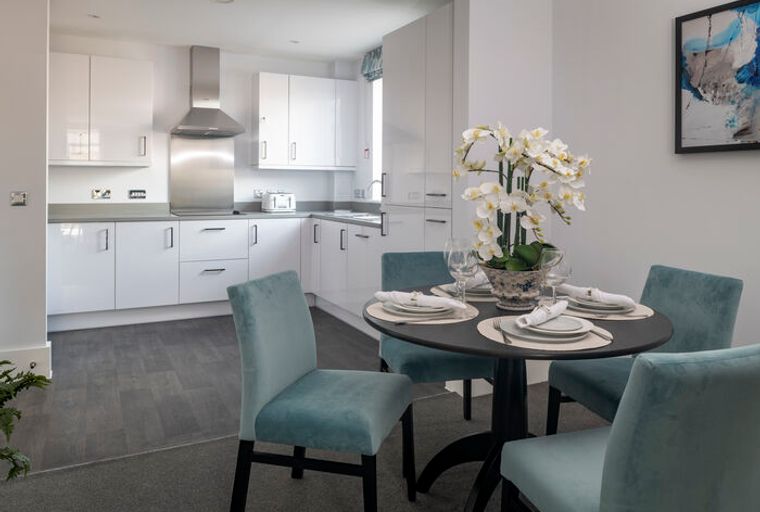 A dining area with a round table set for four, adjacent to a modern kitchen with white cabinets and a stainless steel range hood.