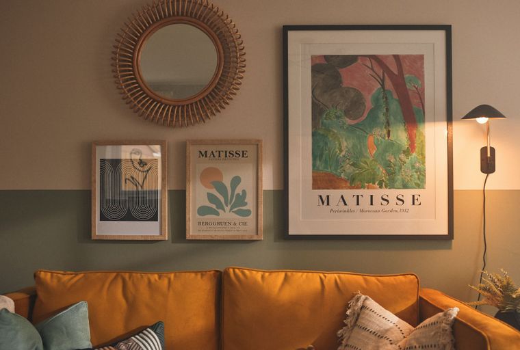 A living room with a mustard yellow sofa, framed artwork, and a rattan mirror on the wall.
