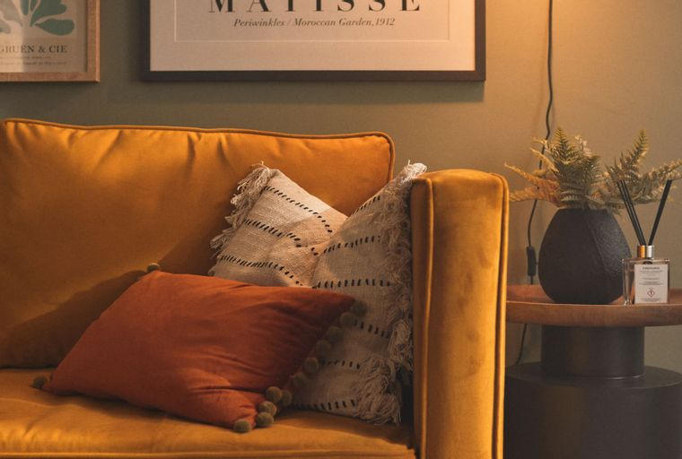 A mustard-yellow sofa with throw pillows and a side table with a vase and diffuser.