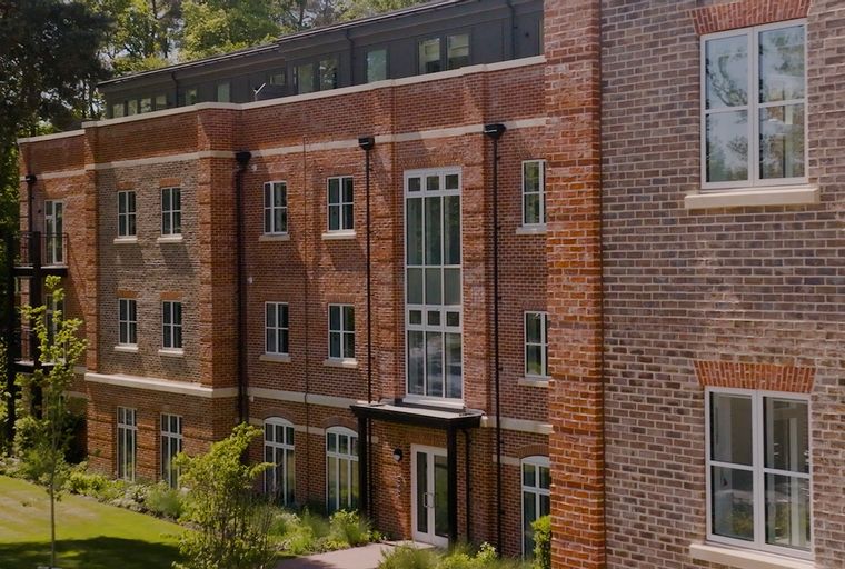 A brick building with multiple windows surrounded by trees and a lawn.
