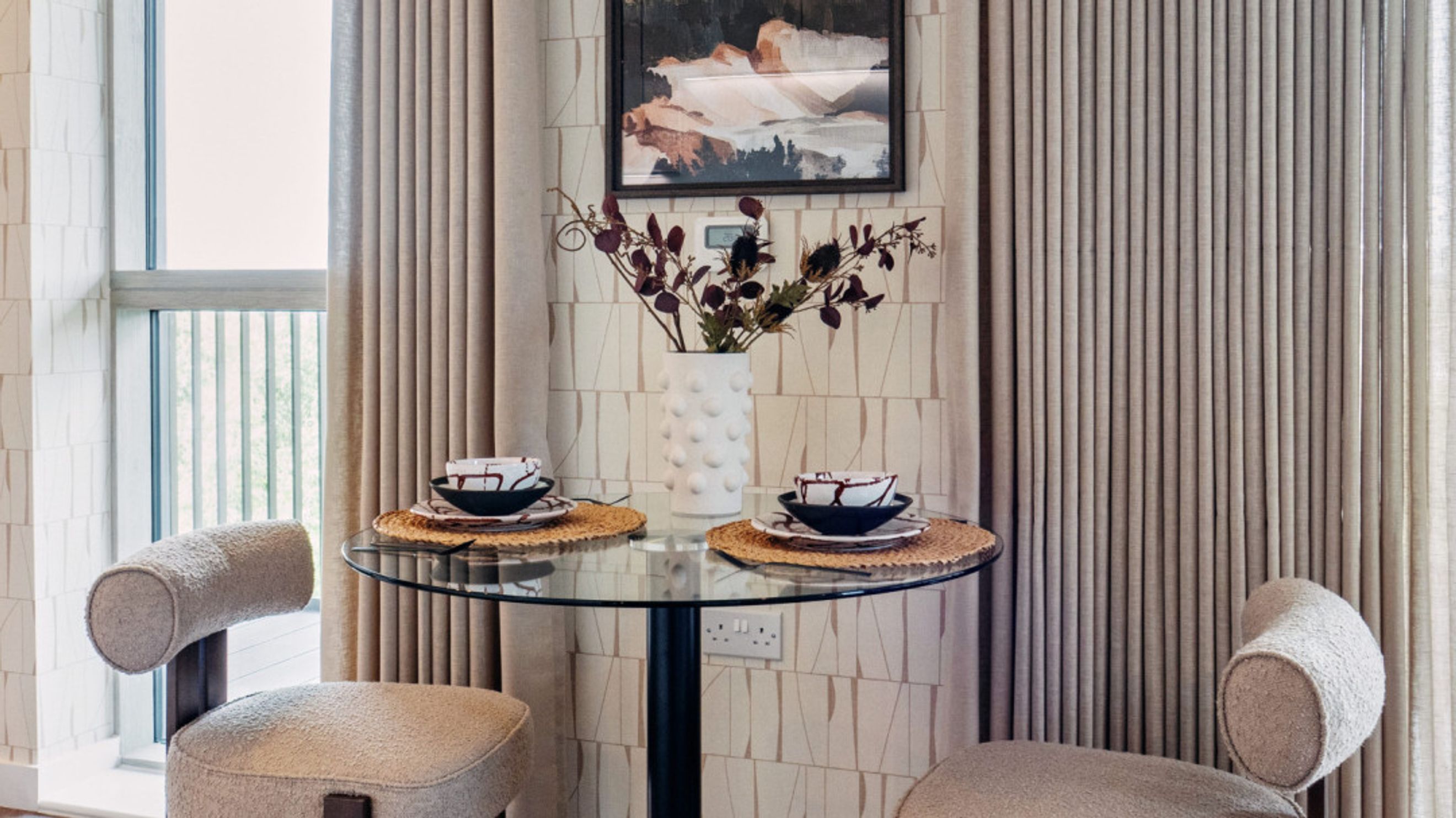 A glass-top dining table with two chairs, a vase of flowers, and a painting on the wall.