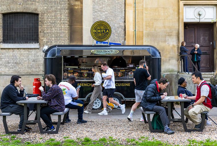 A food truck named BOKIT'LA French Cuisine is parked outside a building, with several people sitting at picnic tables and walking around.