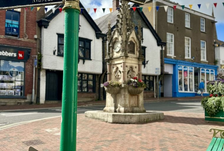 A town square with a monument, shops, and a green lamppost in the foreground.