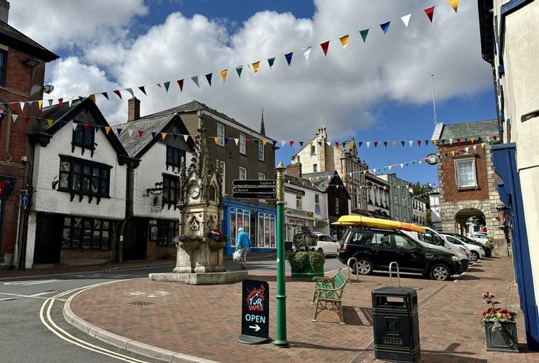 A town square with buildings, bunting, a clock tower, and parked cars.