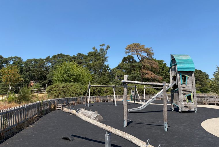 A playground with a wooden playhouse, slide, swings, and a seesaw on a rubberized surface.