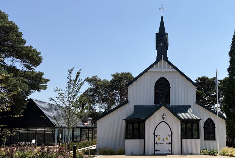A white corrugated iron church with a dark green roof and steeple, set against a blue sky.