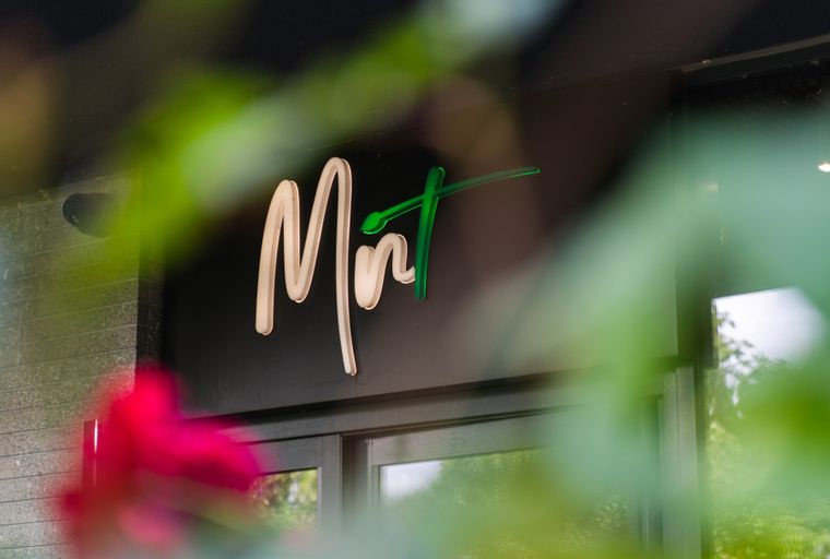 A sign that says 'Mint' is displayed on a dark building facade, partially obscured by green foliage.