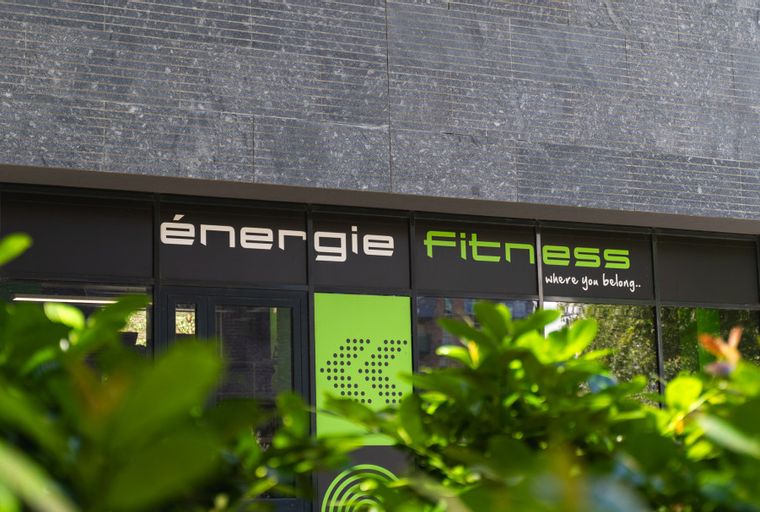 A storefront for Energie Fitness with green and white signage, partially obscured by green leaves.