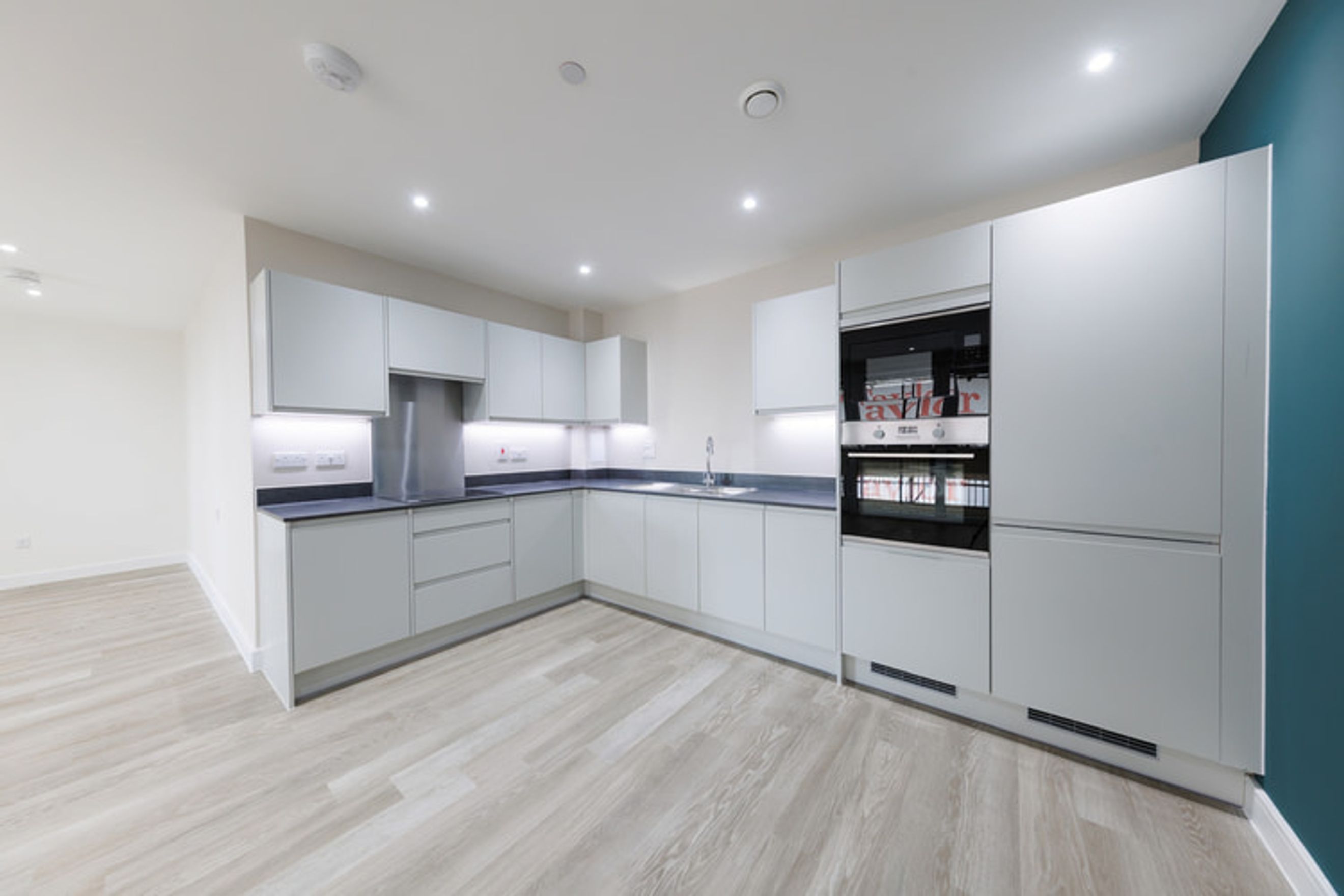 A modern L-shaped kitchen with light grey cabinets, dark countertops, and integrated ovens.