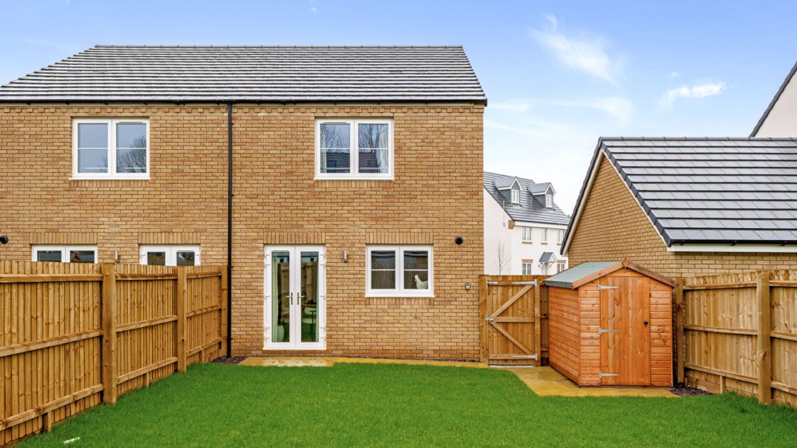 A backyard with a brick house, wooden fence, garden shed, and green lawn.