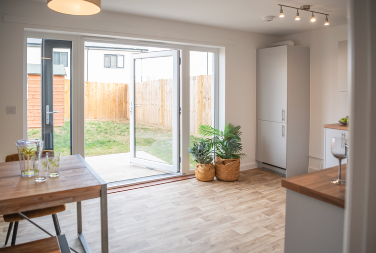 A dining area with a wooden table and chairs, opening to a garden through double doors, with a kitchen area visible.