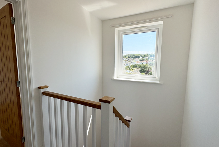 A staircase with a wooden handrail and white balusters next to an open wooden door and a window overlooking a town.