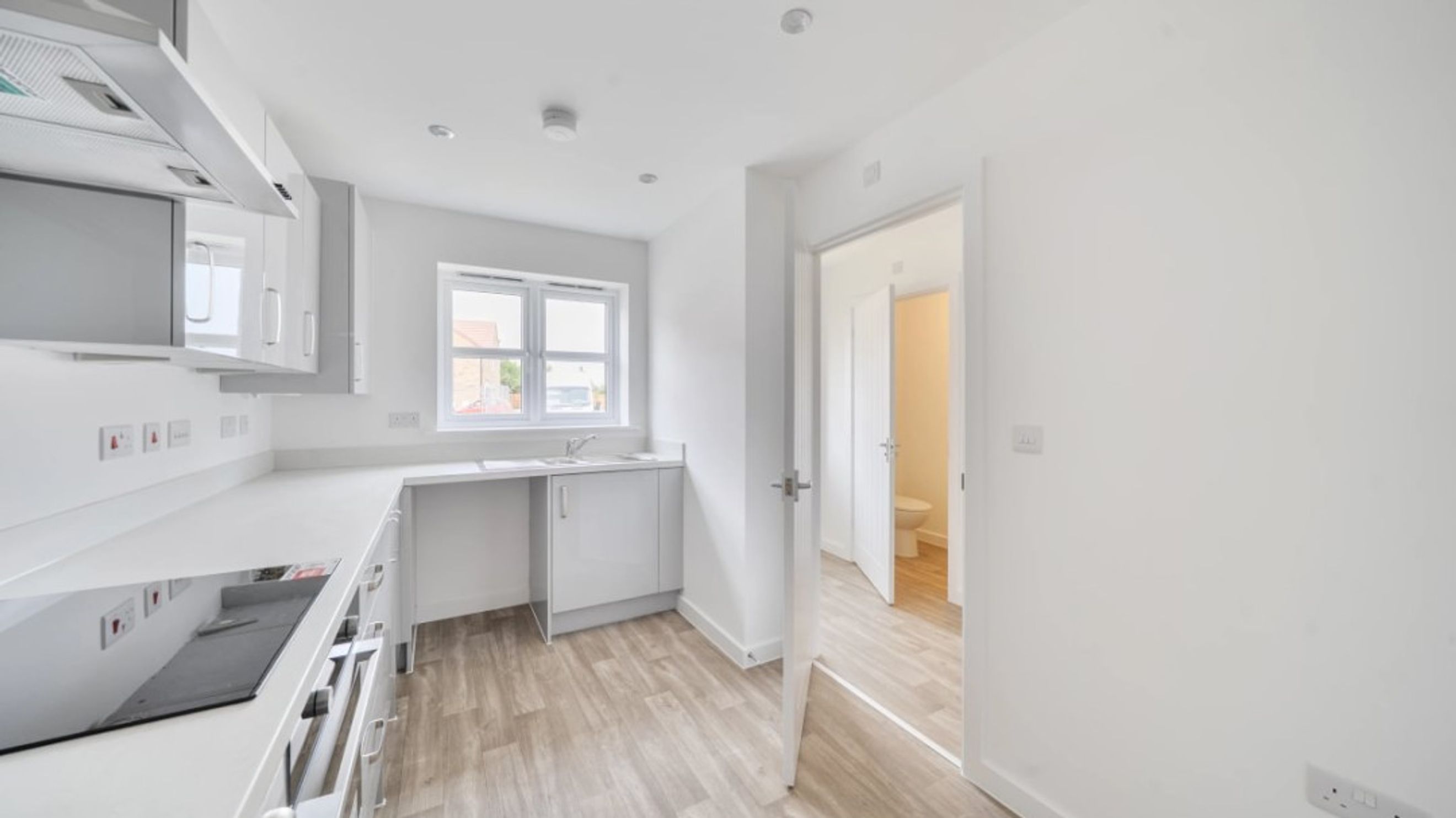 A modern kitchen with white cabinets and countertops, a hob, and a window, with a doorway leading to a bathroom.
