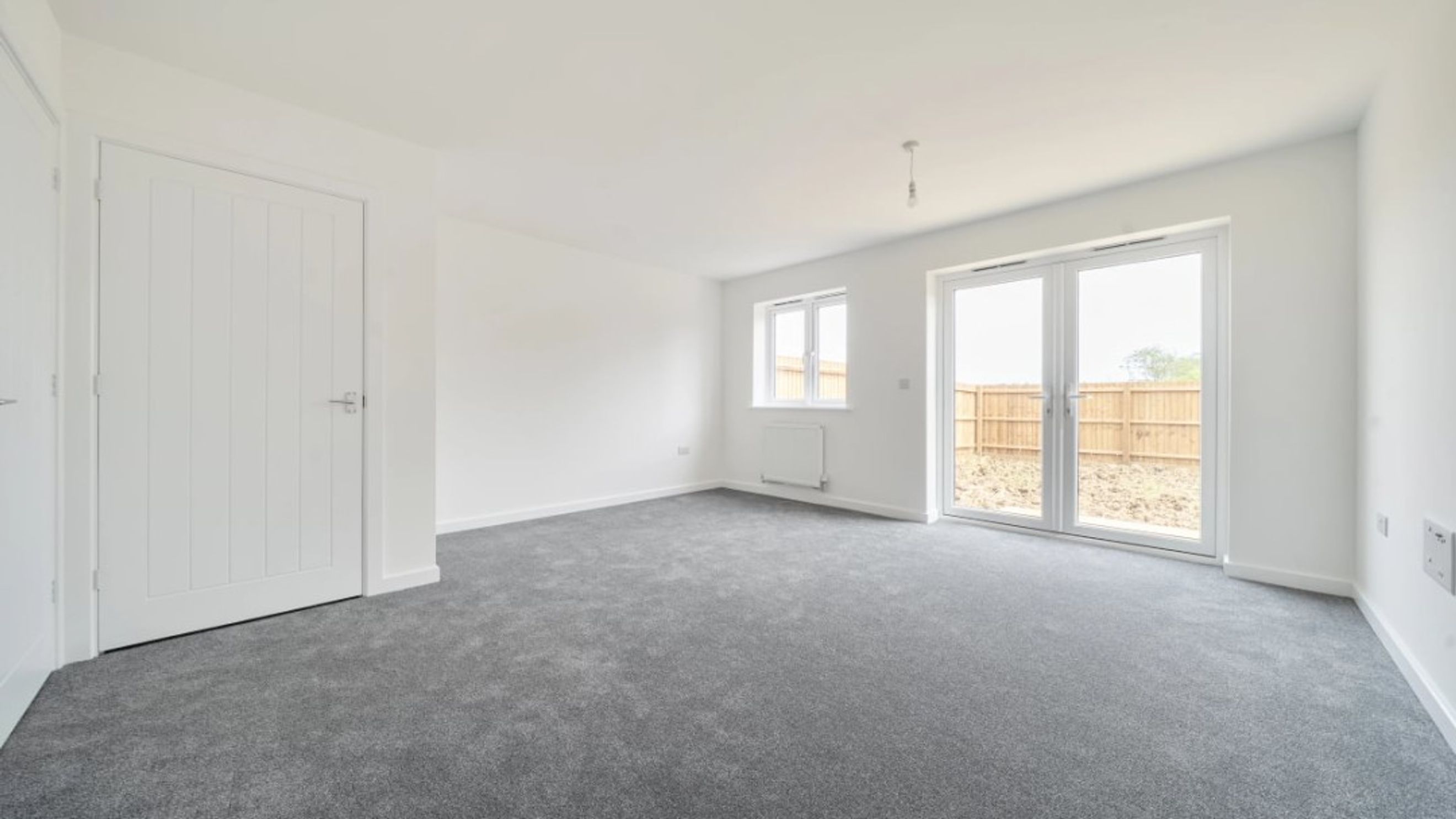 A spacious, unfurnished room with grey carpet, white walls, a white door, a window, and double doors leading outside.