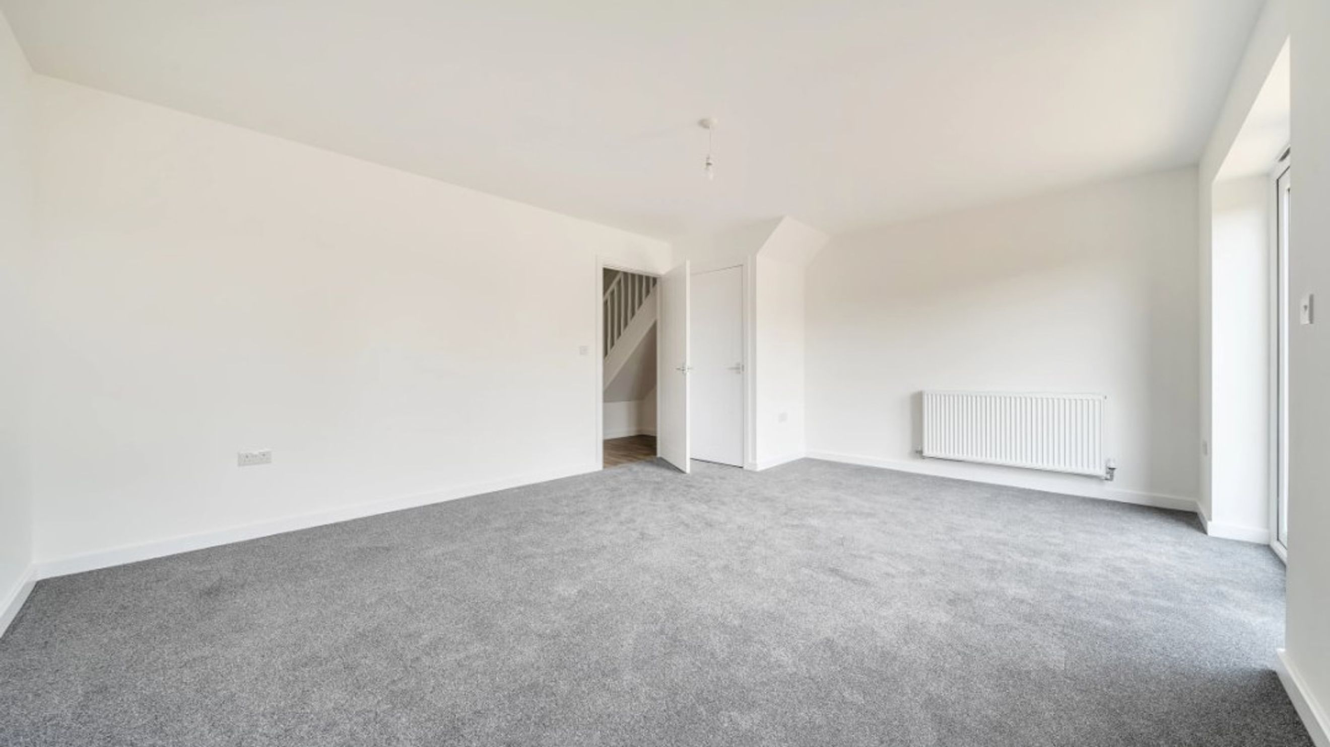 A spacious, empty room with grey carpet, white walls, a radiator, and a doorway leading to stairs.