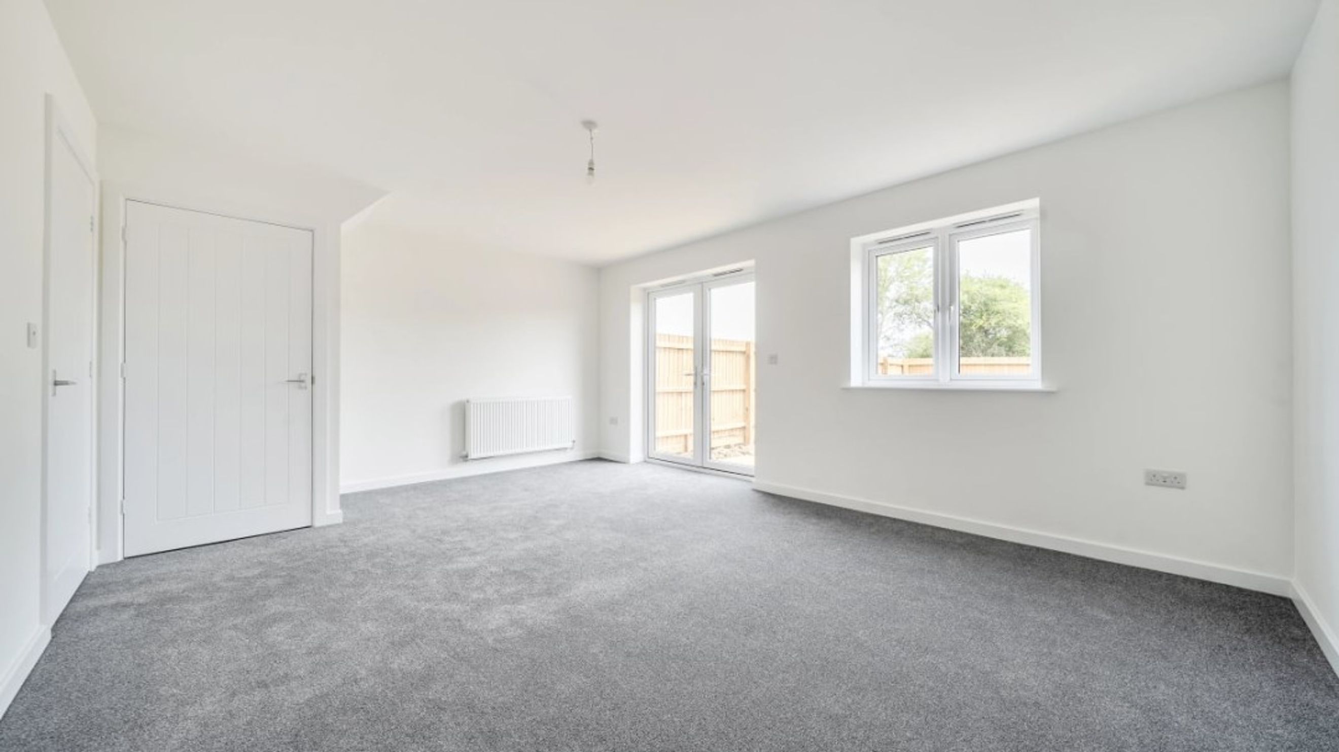 A living room with grey carpet, white walls, a radiator, double doors, and a window.
