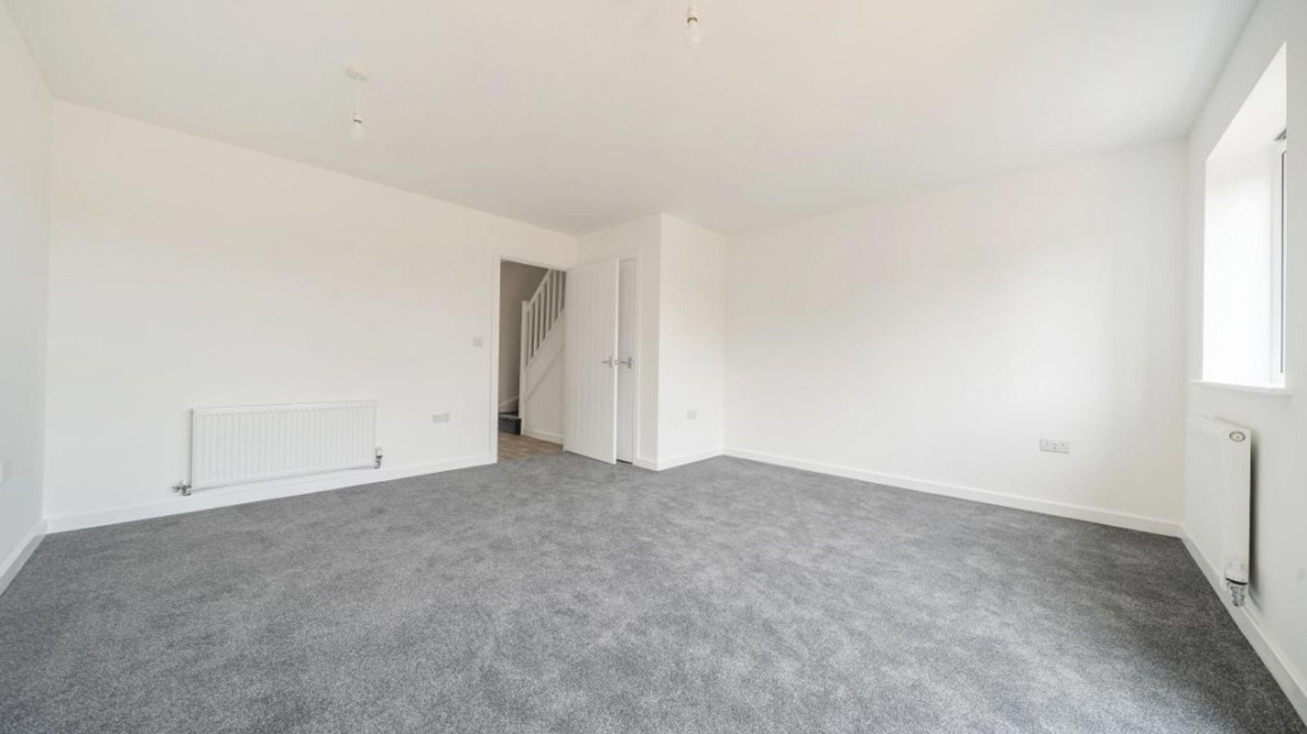 A spacious, unfurnished room with grey carpet, white walls, and a radiator.