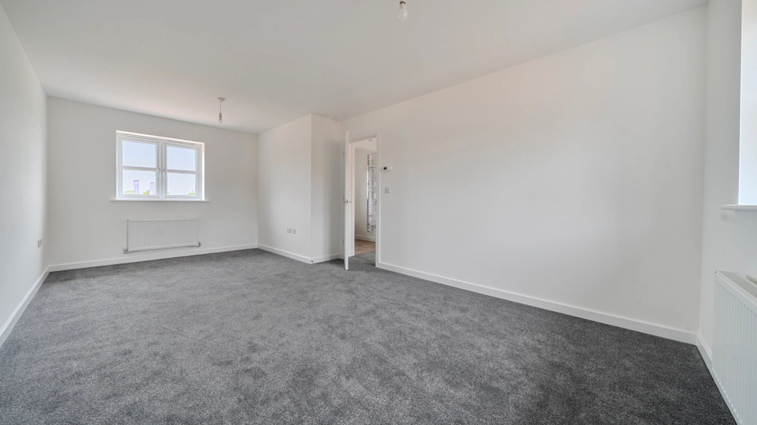 A spacious, empty room with grey carpet, white walls, and a window.