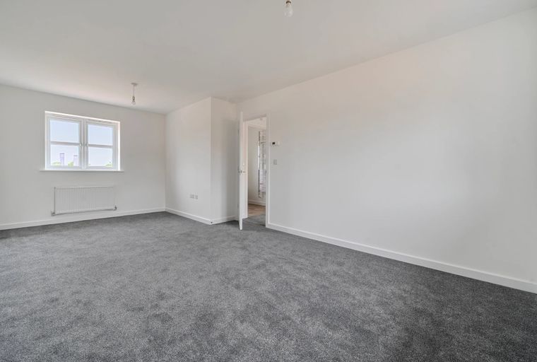 A spacious, empty room with grey carpet, white walls, and a window.