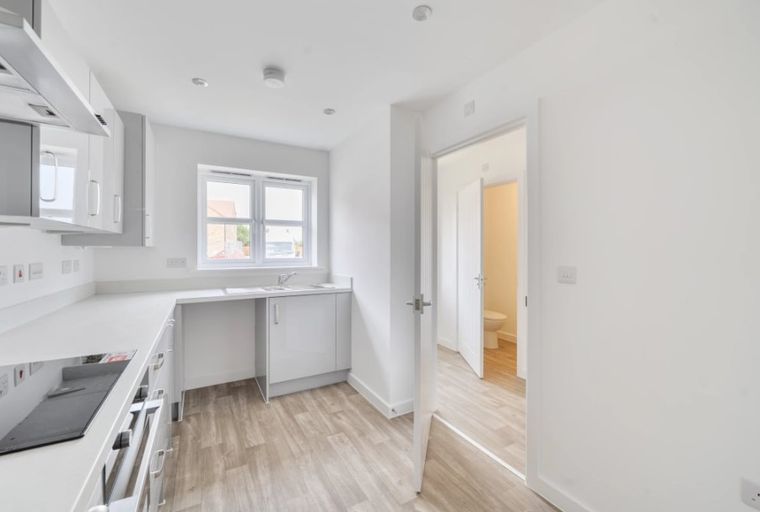 A modern kitchen with white cabinets and countertops, a hob, and a window, with a doorway leading to a bathroom.