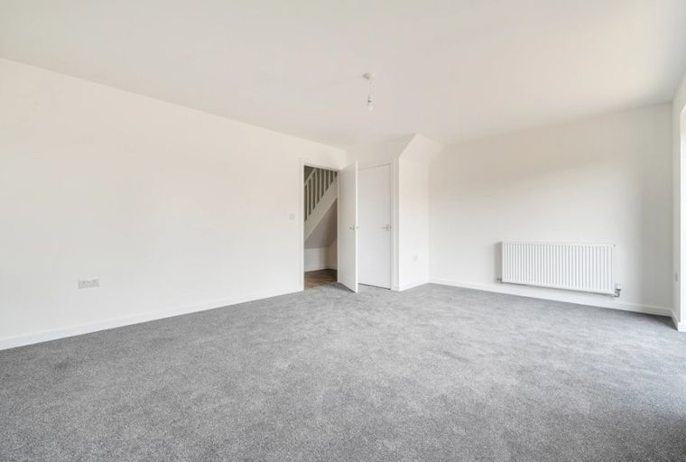 A spacious, empty room with grey carpet, white walls, a radiator, and a doorway leading to stairs.