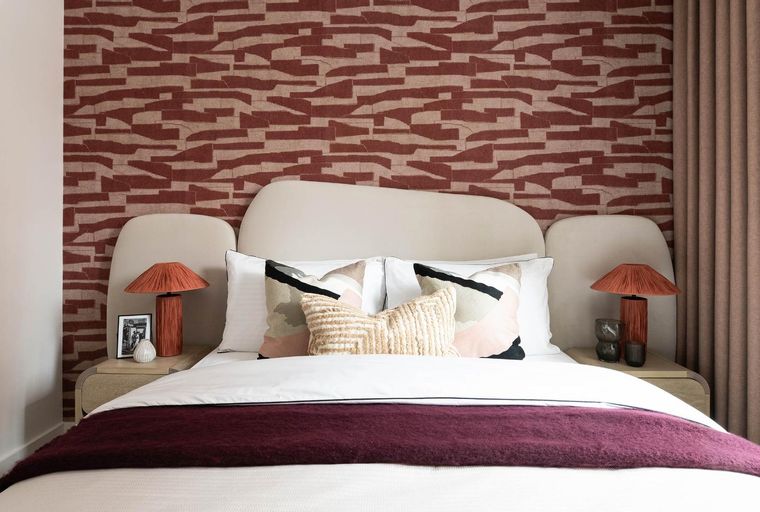 A bedroom with a bed, pillows, lamps, and patterned wallpaper.