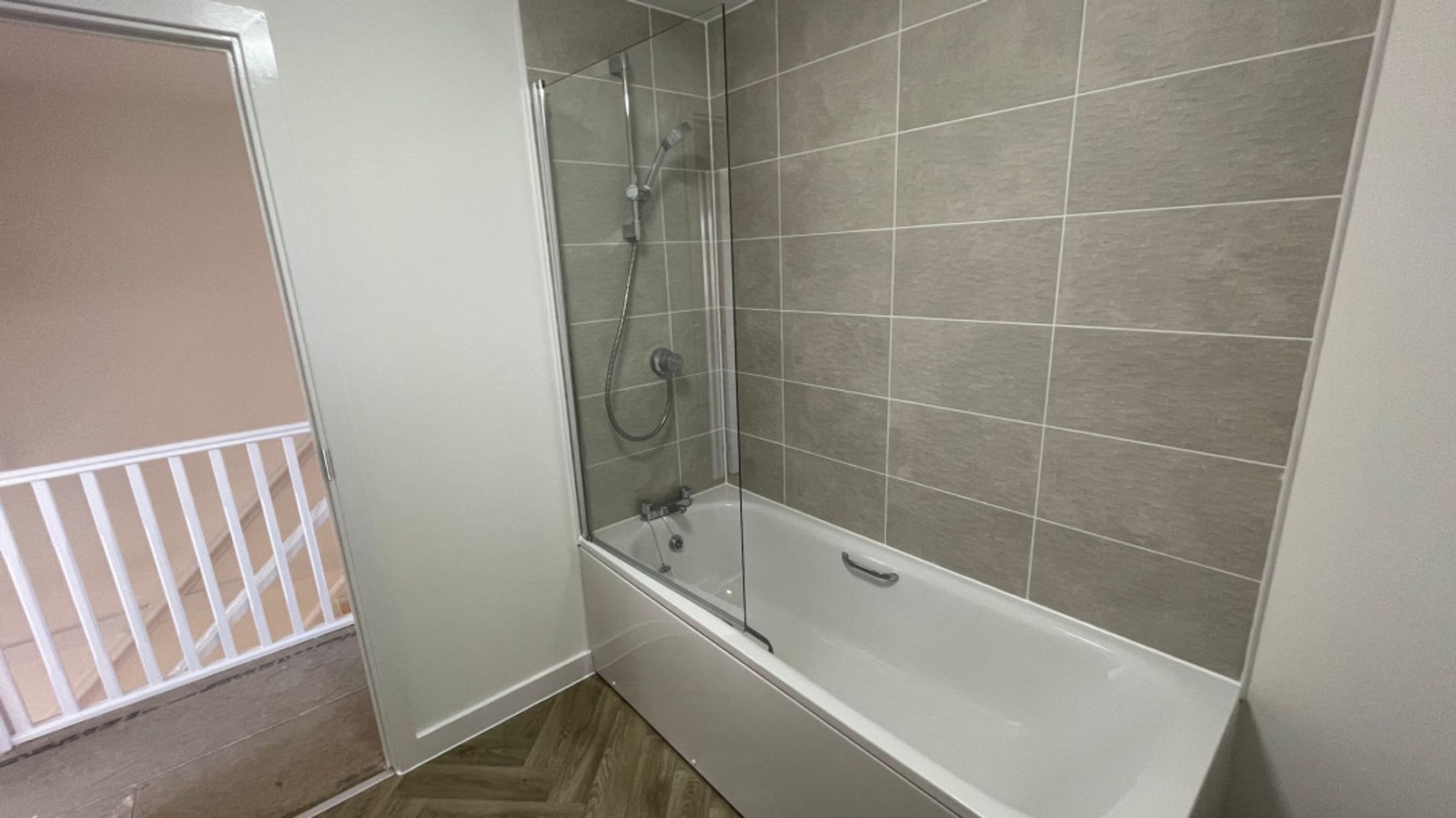 A bathtub with a glass shower screen and grey tiled walls in a bathroom.